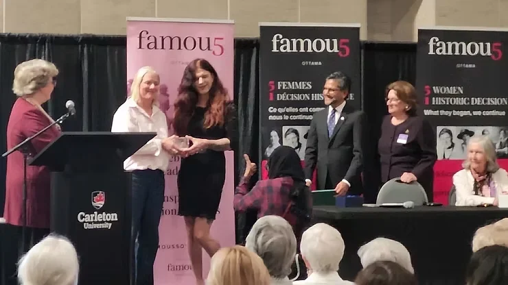 A photograph taken at an indoor awards event on a stage, with a wooden lectern reading "Carleton University" in the foreground on the left, where a woman with short grey hair and glasses stands at a microphone wearing a maroon blazer. Beside her, a woman (Catherine McKenna) with short blonde hair in a white long-sleeve top clasps hands with a man (Zachary "Fae" Johnstone) who has long, wavy dark red hair and wears a black dress. He is several inches taller than McKenna. To their right stand a man in a dark suit and a woman with short reddish hair and glasses wearing a dark blazer with a name badge, both smiling and clapping. In the foreground, partially visible, a person wearing a maroon-and-black plaid outfit and a black hijab crouches or bends near the stage. An older woman with white hair sits at a table on the far right. Behind the group are three pink and black vertical banners reading "famou5, OTTAWA," "5 FEMMES, 1 DÉCISION HIS[TORIQUE]," and "5 WOMEN, 1 HISTORIC DECISION" with black-and-white photos of women. A seated audience is visible in the foreground, out of focus.