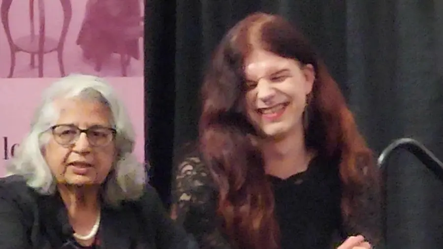 A close-up photograph of two people seated on a stage in front of a dark curtain, with a pink "famou5" banner partially visible in the background on the left, along with a microphone stand on the right. On the left, an older woman (Dr. Indu Gambhir) with wavy white hair, glasses, and a pearl necklace wears a dark top and looks off to the side. Beside her, a man (Zachary "Fae" Johnstone) with long, wavy dark red hair parted in the middle is smiling broadly with mouth open and nose scruched, wearing a black lace-patterned top. His adam's apple is clearly visible.