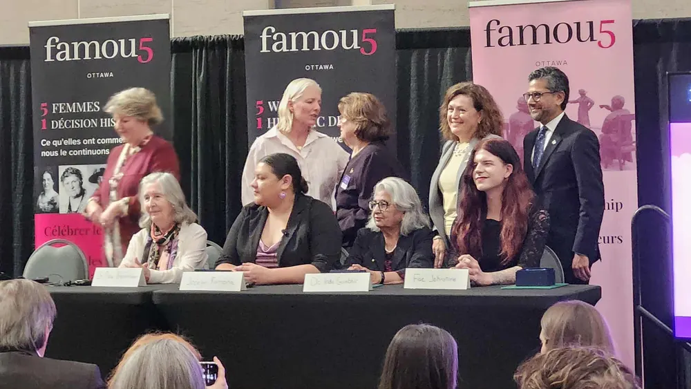 A photograph taken on an event stage with three pink and black "famou5, OTTAWA" banners visible in the background, one reading "5 WOMEN, 1 HISTORIC DECISION, What they began, we continue" with black-and-white photos of women. On stage, a row of people stands and sits behind a black-draped table with white name placards. Standing in back, from left to right: a woman with white hair partially obscured mid-motion, a woman with long blonde hair in a white top, an older woman in a dark cardigan, a woman with curly reddish-brown hair in a beige blazer, and a man in a dark suit. Seated at the table, from left to right: an older woman with grey hair in a light cardigan (nameplate partially reads "Dr. Indu Gambhir" or similar), a woman with dark hair pulled back wearing a purple top and black cardigan, an older woman with grey hair in a black top, and a man (Zachary "Fae" Johnstone) with long, wavy dark red hair wearing a black outfit, smiling with hand resting on the table. A seated audience is visible in the foreground, out of focus.