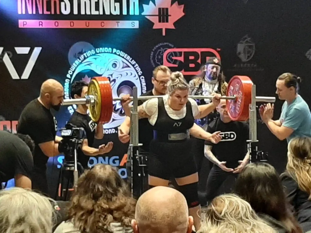 A powerlifting competition photo showing a woman with light brown hair in a bun performing a barbell squat, wearing a black singlet with an "A7" logo over a white long-sleeve top, a weightlifting belt, and knee sleeves. The barbell is loaded with large red plates on each side. Several spotters and officials in black shirts (one branded "SBD") stand close around her ready to assist, with a referee at right giving a hand signal. A cameraman films from the left, and an audience of spectators is visible in the foreground. The backdrop is a black step-and-repeat banner featuring sponsor logos including "InnerStrength Products," "A7," "SBD," and a "Canadian Powerlifting Union" emblem with a stylized eagle/thunderbird design.