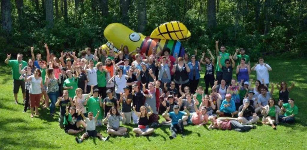 An outdoor group photo on a grassy field in front of a dense treeline, taken in daylight. A large crowd of roughly 60-70 people of mixed ages stands and sits in a loose formation, most making a "rock on"/horns hand gesture with one or both hands raised. Many wear green T-shirts (some with logos), while others wear a mix of casual outfits in black, white, and colorful patterns. In the center background stands a large inflatable mascot/balloon figure with a yellow bulbous head, large white cartoon eyes, a wide grin, and a body striped in rainbow colors (red, orange, yellow, green, blue, purple). Several children sit cross-legged in the front row on the grass.