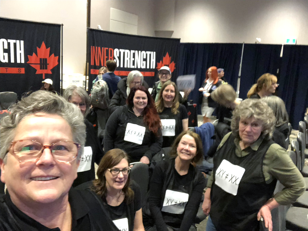 A selfie taken by Linda Blade in the foreground, wearing glasses and a black top, smiling at the camera. Behind her, a group of roughly ten women, all dressed in black, sit and stand together, each wearing a printed "XY ≠ XX" sign pinned or taped to their chest. Several are smiling for the photo. The setting matches the same indoor event venue seen in the earlier photos, with "InnerStrength Products" banner signage (featuring a red maple leaf) visible in the background, along with other event attendees and stacked chairs.