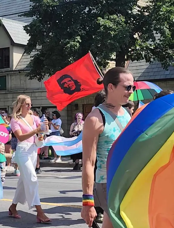 A photograph taken at an Ottawa pride parade 2024 on a sunny day. A man with a low ponytail, sunglasses, and a neck tattoo walks in the foreground wearing a tie-dye tank top with a rainbow-striped wristband, carrying or standing near an inflatable rainbow-colored object. Behind him, a red flag featuring a black stylized illustration of a man's face wearing a beret with a star (the iconic Che Guevara image) flies on a pole, with small illegible text visible at the bottom of the flag. To the left, a woman in a pink top and white pants holds a water bottle, and other parade participants and a stone building with dark windows are visible in the background alongside a rainbow-colored umbrella.