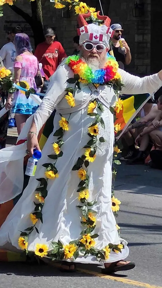 A man wearing a white dress and a light pink chest harness with a rainbow lei around his neck and artificial sunflowers draped over his sholders. He has a floral headpiece combined with Canadian-flag-themed elements (red and white, maple leaf motifs) at Ottawa Pride 2024.