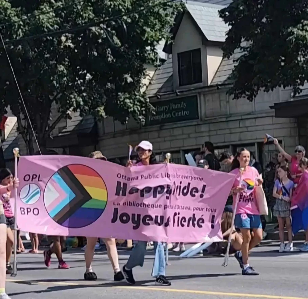 A photograph of a parade scene showing several people carrying a large pink fabric banner suspended between poles down a city street, in front of a stone building with a visible sign reading "...arry Family Reception Centre" and green trees overhead. The banner reads, in dark purple text: "OPL BPO" beside a circular Progress Pride flag logo (rainbow stripes with a chevron of white, pink, light blue, brown, and black pointing into the circle), followed by "Ottawa Public Library for everyone," then in large bold script "Happy Pride!" with French text below reading "La bibliothèque pour d'Ottawa, pour tous," and beneath that, "Joyeuse fierté!" People in the parade wear pink T-shirts, and one visible shirt appears to read "...e Fierté" (Pride). Onlookers stand along the sidewalk, some with rainbow-colored items visible.