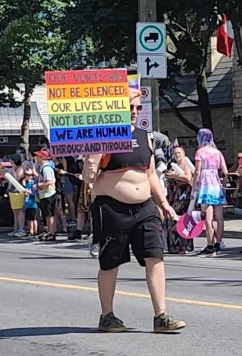A handheld sign reading "OUR LIVES WILL NOT BE ERASED" in bold capital lettering, held up by a woman in a crop top and cargo shorts at Ottawa Pride 2024