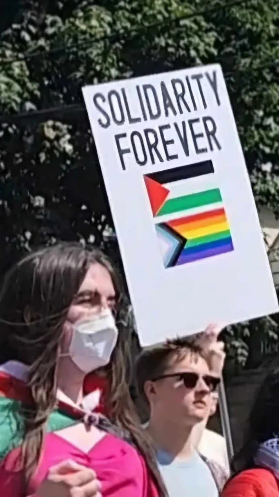 A sign or banner reading "SOLIDARITY FOREVER" with additional smaller text referencing "QUEER & TRANS FOR PALESTINE", people are in the foreground, one is wearing a white surgical mask with the palestine flag tied around their neck.