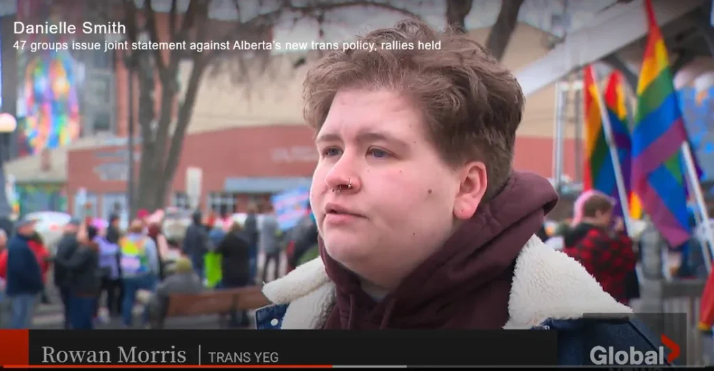 A screenshot of a television news broadcast. In the upper left, white on-screen text identifies the speaker as "Danielle Smith" with a headline below reading "47 groups issue joint statement against Alberta's new trans policy, rallies held." The main frame shows a close-up of a woman (Rowan Morris/Venedam) with short curly reddish-brown hair, a nose piercing, wearing a maroon hoodie under a denim jacket with a fleece collar, looking off to the side while speaking, standing outdoors at a rally with a blurred crowd and multiple large rainbow and transgender pride flags visible in the background, along with string lights and colorful building murals. At the bottom of the screen, a black lower-third graphic identifies the woman as "Rowan Morris" with the label "TRANS YEG" beside her name, and the "Global" news network logo with a red arrow icon appears in the bottom right corner. A thin red progress/scrub bar runs along the very bottom edge of the video frame.