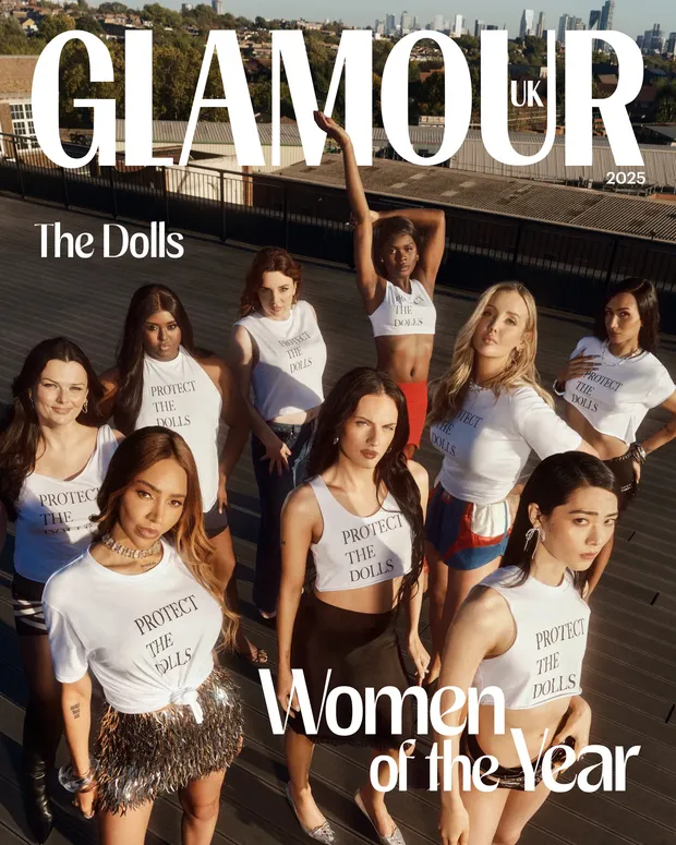 A magazine cover for "GLAMOUR UK, 2025." Large white bold text at the top reads "GLAMOUR" with "UK" in smaller text beside it. Below, on the left, italicized white text reads "The Dolls." The cover photo shows nine men posed on a rooftop with a city skyline in the background, all wearing white cropped T-shirts or tank tops printed with the black text "PROTECT THE DOLLS," styled with different bottoms (jeans, shorts, skirts). One man in the back has his arm raised. At the bottom of the cover, large white text reads "Women of the Year."