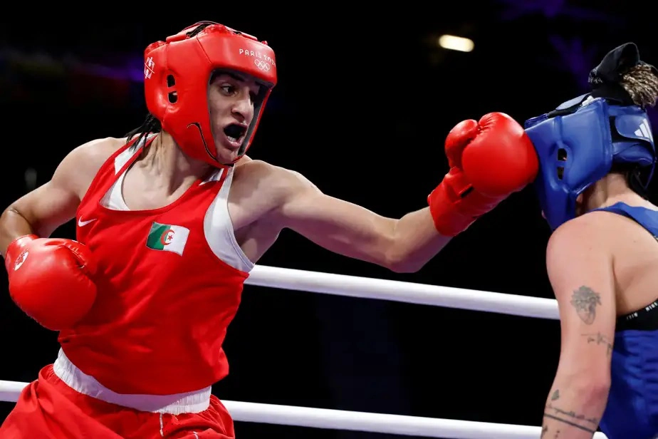 An action shot from an Olympic boxing match (Paris 2024). Imane Khelif in red gear with an Algerian flag patch on his jersey throws a right-hand punch that connects with his opponent's blue headgear. The opponent, Luca Hamori, wearing blue gear with a visible tattoo on her upper arm, is caught mid-impact. Both are inside a boxing ring with white ropes visible in the background.