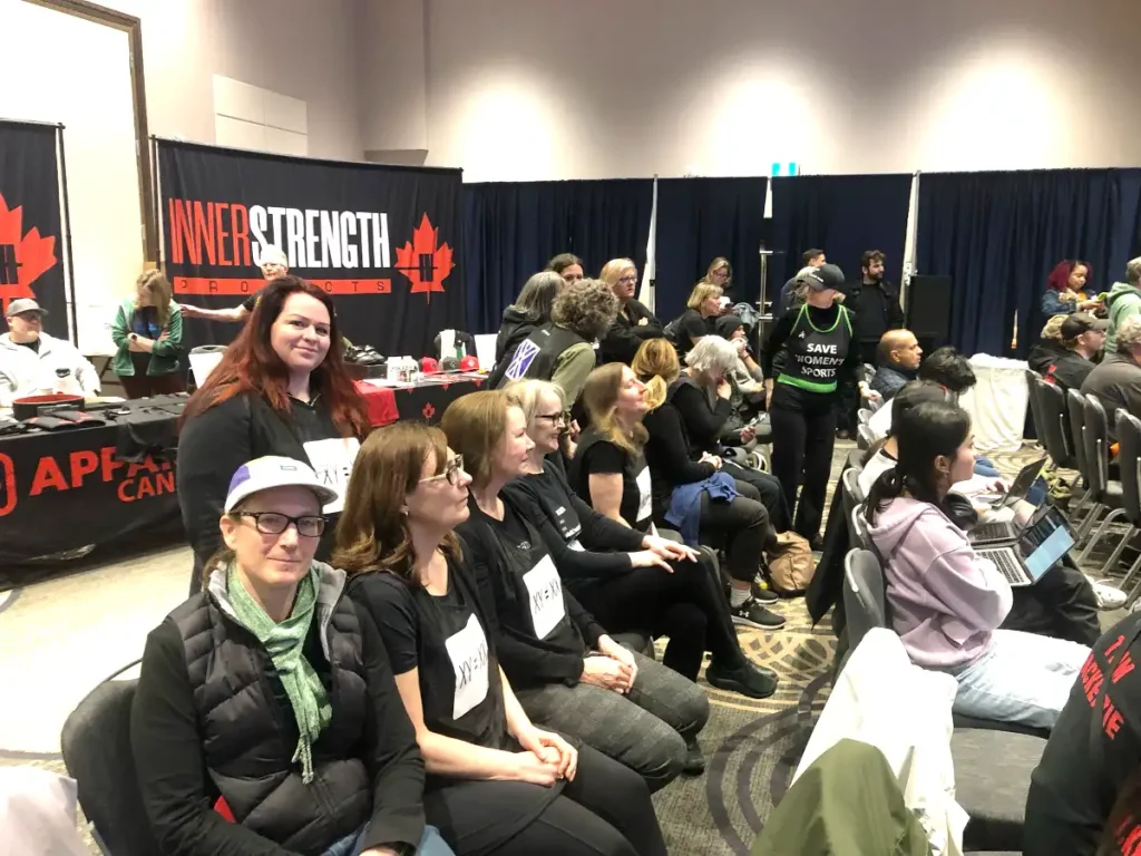 A group of women seated in rows at the same indoor event, several wearing matching black t-shirts with a printed "XX = XX" design on the chest (a reference to female chromosomes) and paper name-tag-style signs pinned to their shirts. One woman stands behind them in a black jacket, smiling at the camera, near a merchandise table. In the background, a person wears a black tank top reading "SAVE WOMEN'S SPORTS" in green lettering, and another visible sign fragment reads "1 W...ONE TIME." A vendor booth backdrop reads "InnerStrength Products" with a red maple leaf design, and additional attendees and tables are visible further back in the room.