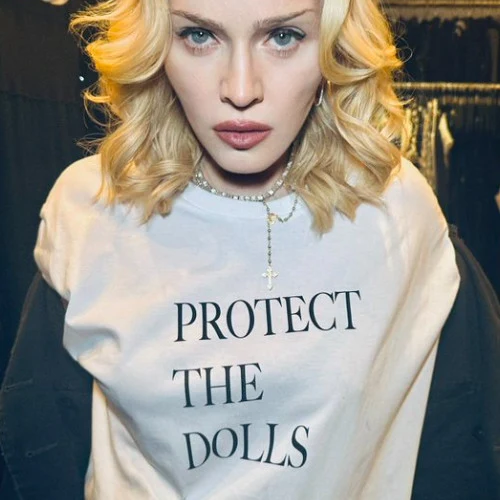 A close-up photograph of pop singer Madonna with curly platinum blonde shoulder-length hair, pale makeup, and pink lipstick, looking directly at the camera with a serious expression. She wears a silver rosary-style necklace with a cross pendant and a white long-sleeve T-shirt printed with black text reading "PROTECT / THE / DOLLS," stacked in three lines. She stands indoors in a dim setting with dark clothing racks visible blurred in the background.