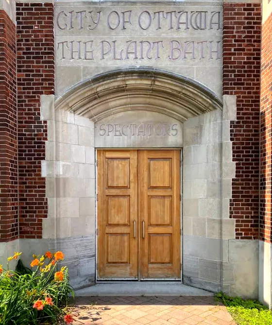A photograph of an ornate stone building entrance with red brick pillars flanking a carved limestone archway. Carved into the stone above the arch, in an aged, weathered font, reads "CITY OF OTTAWA / THE PLANT BATH," and directly above the wooden doors within the arch, smaller carved text reads "SPECTATORS." Below this, a pair of modern wooden double doors with vertical panel detailing and metal handles serves as the entrance. Orange daylily flowers bloom in a garden bed at the bottom left of the frame.