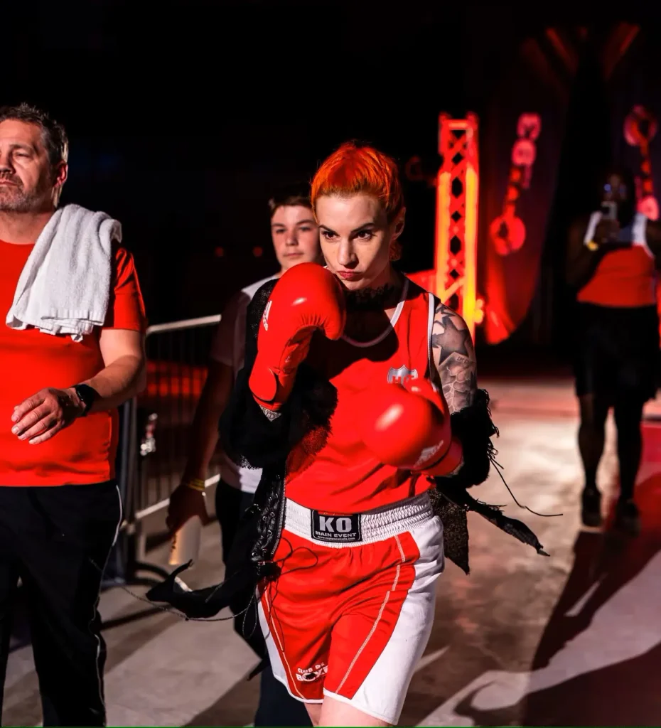 A dramatic action photo of a boxer entering an arena, wearing a red boxing jersey with a shield logo, white-and-red shorts with a "KO Main Event" waistband, red boxing gloves raised in a guard position, and short bright orange hair. She has visible tattoos on one shoulder/arm and a neck tattoo, with a dark shawl-like garment draped over one shoulder. Behind her, a man in a red shirt with a towel over his shoulder and a teenager in a white shirt walk alongside her; red-lit stage scaffolding and decorative elements are visible in the background, suggesting a themed boxing event entrance.
