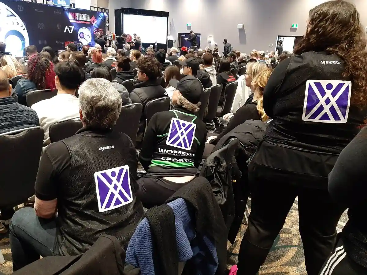 A crowd of spectators seated (and some standing) at an indoor event, viewed from behind, watching a stage in the distance where a powerlifting competition taking place (visible barbell setup and "InnerStrength Products"/"A7"/"SBD" sponsor branding on the backdrop, matching the previous powerlifting photo). Several people in the foreground wear matching black jackets with a purple-and-white geometric "X" logo featuring a stick-figure person on the back; one jacket partially reads "WOMEN'S SPORTS." The jackets have the logo of the ICFS on them.