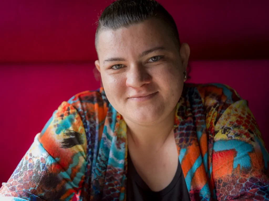 A close-up portrait photograph of a female detransitioner with short, closely cropped dark hair, an ear piercing, and a slight, warm smile, looking directly at the camera. She is wearing a colorful, patterned jacket in shades of orange, teal, yellow, and dark blue over a black top. The background is a solid deep magenta/red, with soft studio-style lighting on her face.