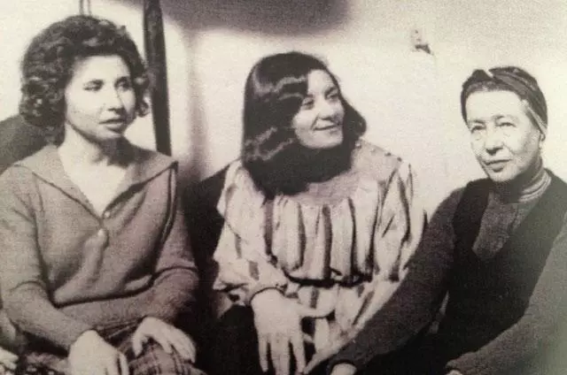 An old black-and-white photo of three women seated closely together. The woman on the left has curly dark hair and wears a grey cardigan; the woman in the middle has voluminous curly dark hair and wears a patterned ruffled blouse; the woman on the right has short grey hair pulled back with a headscarf/band and wears a dark top over a striped underlayer.