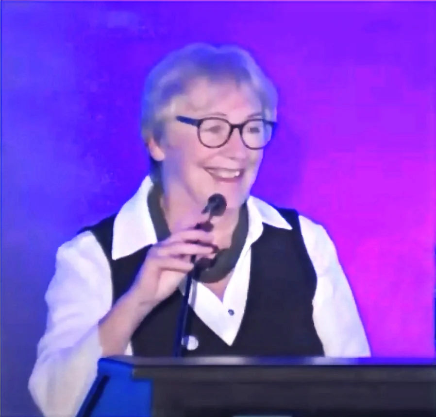 A photo of Ghislaine Gendron speaking at a podium with a microphone, against a stage lit with purple and blue lighting. She has short grey/white hair, wears dark-rimmed glasses, a black vest over a white collared shirt, and a grey scarf around her neck. She's smiling broadly mid-sentence, with one hand raised near the microphone.