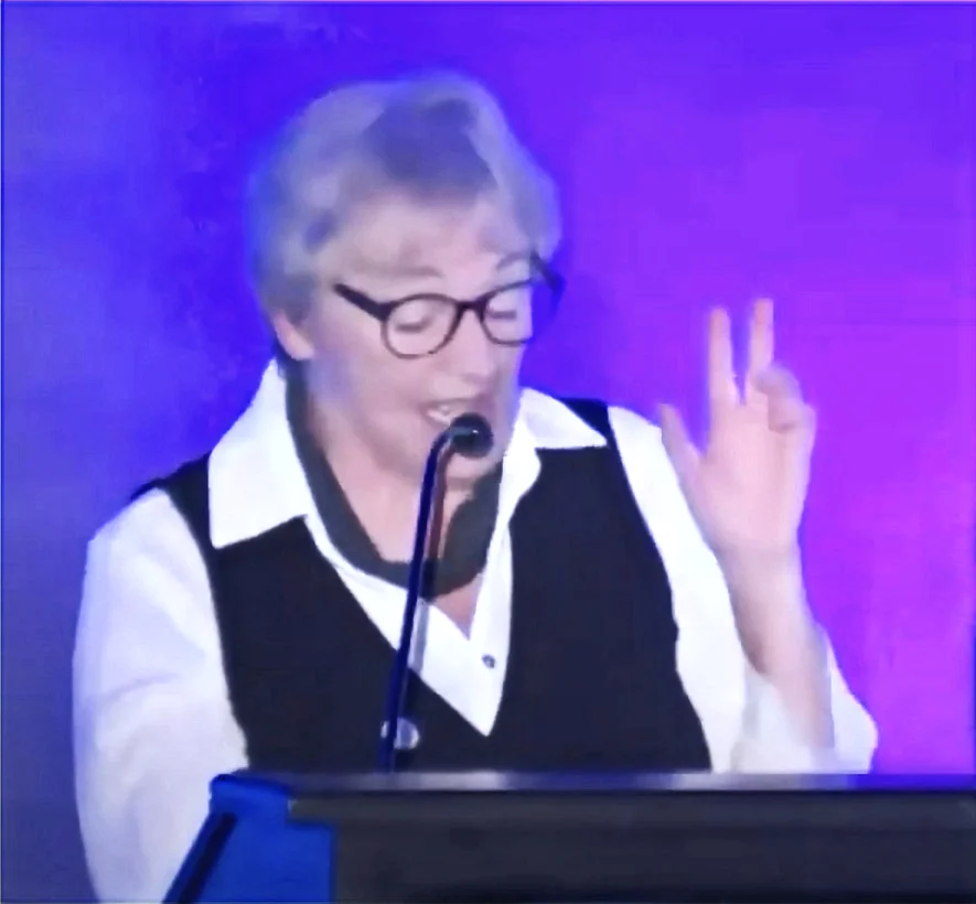 A photo of Ghislaine Gendron speaking at a podium with a microphone, against a stage lit with purple and blue lighting. She has short grey/white hair, wears dark-rimmed glasses, a black vest over a white collared shirt, and a grey scarf around her neck. She appears to be laughing, with her eyes closed and one hand raised.