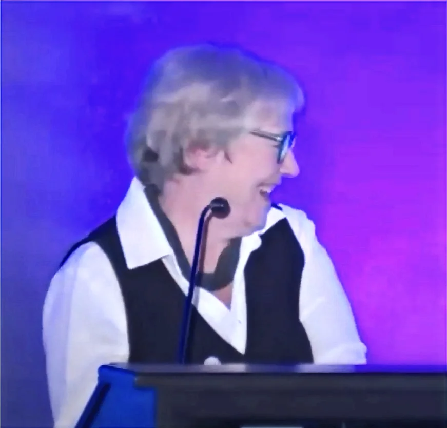A photo of Ghislaine Gendron speaking at a podium with a microphone, against a stage lit with purple and blue lighting, shown in left profile. She has short grey/white hair, wears dark-rimmed glasses, a black vest over a white collared shirt, and a grey scarf around her neck. She is laughing with her mouth open and her head tilted back slightly.