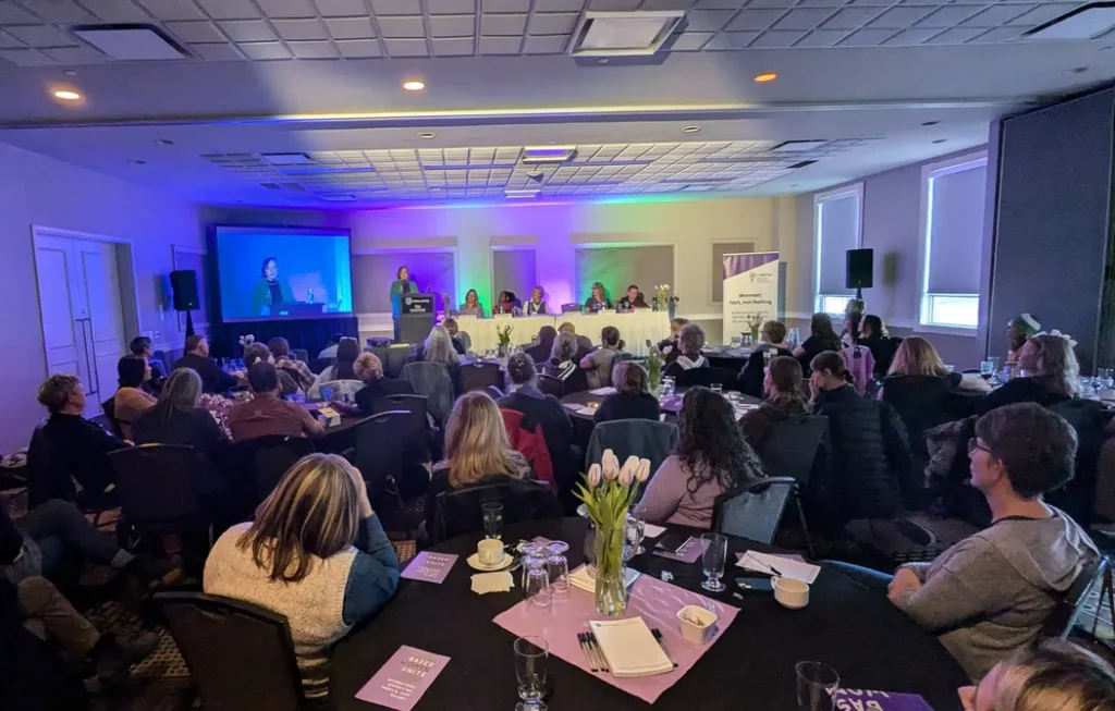A wide photo of a conference room at a hotel, with a purple and green colored stage wash lighting the front wall. A woman stands at a podium speaking, with a large projector screen beside her showing a livestream of the event. A panel of five seated women faces the audience at a long white-clothed table behind the podium, with a "caWsbar" banner reading "Woman: fact, not feeling" to the side. In the foreground, a full room of seated attendees at round tables covered in black cloths with pink placemats, glassware, tulip centerpieces, and small purple cards reading "REALITY BASED WOMEN UNITE" and "INTERNATIONAL WOMEN'S DAY / MARCH 8, 2025 / CALGARY."