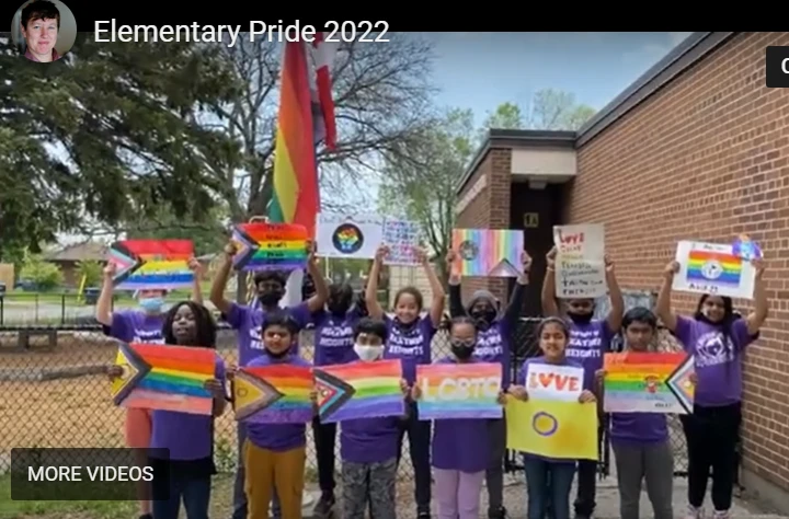 A paused video frame titled "Elementary Pride 2022" (with a small circular profile photo of a woman in the top-left corner and a "MORE VIDEOS" label in the bottom-left corner). It shows a row of children outside a brick school building, several wearing purple T-shirts and face masks, holding up handmade signs and pride flags — including a rainbow flag with black/brown stripes and the trans-flag chevron (the "Progress Pride" design), a sign reading "LGBTQ+", another reading "LOVE" with a heart-shaped "O", and additional rainbow-colored signs with partially visible text. A large rainbow flag and a red flag with a maple leaf (Canadian flag) fly on poles behind the group.