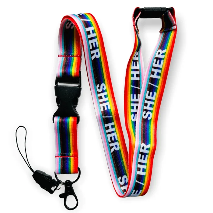 A product photograph of a rainbow-striped fabric lanyard against a white background, arranged in a loose "V" shape. The lanyard has horizontal stripes in the colors of the Progressive Pride flag (black, brown, light blue, pink, white, red, orange, yellow, green, blue, purple) running its length, with the white bold text "SHE/HER" repeated multiple times along the strap. It has a black plastic breakaway clasp near the neck loop and a black plastic swivel hook clip with a clear plastic ID-card loop attachment at the bottom.