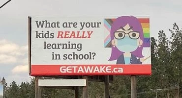 A roadside billboard, photographed from a car window with a highway and treeline visible around it. On a white background, bold black text reads "What are your kids REALLY learning in school?" (with "REALLY" in red italics), next to a cartoon illustration of a woman with purple hair, glasses, and a blue surgical mask, gesturing with one hand, set against a rainbow-striped background. A red banner across the bottom reads "GETAWAKE.ca".