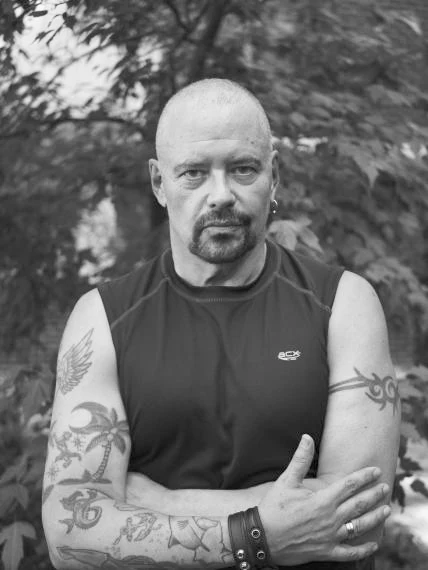 A black-and-white portrait photograph of a bald man with a goatee, standing outdoors in front of leafy foliage. He wears a dark sleeveless athletic top and has his arms crossed, revealing detailed tattoos on both forearms and upper arms, including floral, tribal, and figurative designs, along with a leather wristband and a ring. He looks directly at the camera with a neutral expression.