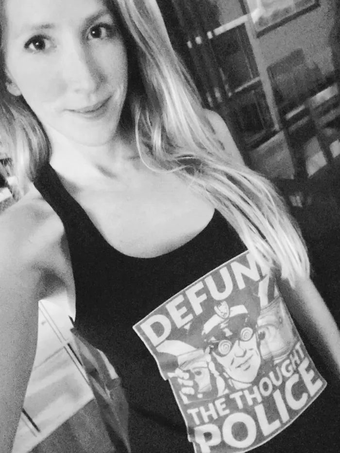 A black-and-white close-up selfie photograph of a woman with long straight blonde hair, smiling slightly at the camera. She wears a black tank top printed with a graphic design featuring an illustrated cartoon police officer with spiral eyes, and text reading "DEFUND" at the top, then "THE THOUGHT" and "POLICE" in bold block letters below. 