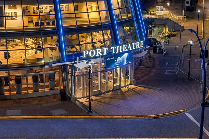 A nighttime photograph of the Port Theatre in Nanaimo, BC with a curved glass facade and prominent angled blue-lit support beams/pillars extending diagonally across the front of the building. Illuminated white signage on a blue background reads "PORT THEATRE" across the entrance canopy. Below the sign, a smaller digital display reads "WELCOME! THE PORT THEATRE" with a ship/anchor logo and additional small text. Through the glass windows, the illuminated lobby interior is visible, including framed photos on the walls and hanging fish sculptures/decorations. Streetlights, a "Museum Way" street sign, and an empty street with a marked crosswalk are visible in the foreground.
