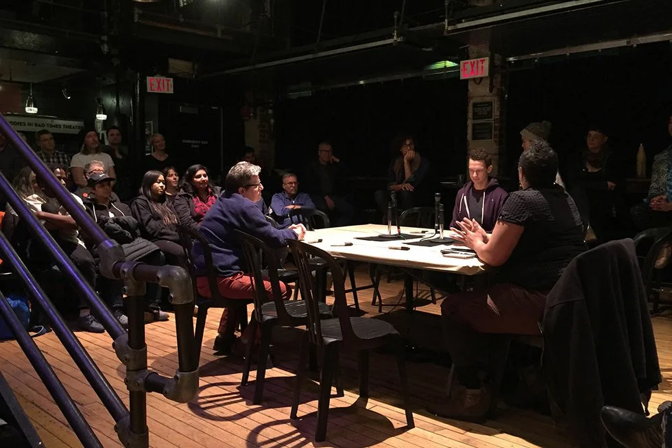 A photograph of an indoor discussion event or panel taking place on a small stage area with exposed pipe railings and string lights hanging from the ceiling. A rectangular table with a white tablecloth is set up with microphones, water bottles, and glasses, around which several people sit facing each other in dialogue, including a person in a blue sweater and a person in dark clothing at the near end, with two more people at the far end of the table. Additional audience members sit and stand around the perimeter of the room in stadium-style seating and along the sides, watching the discussion. Signage reading "EXIT" in red is visible on the wall in the background, along with a partially visible sign reading "... IN BAD TIMES THEATRE" and "EMERGENCY EXIT ONLY."