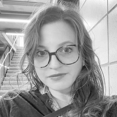A black-and-white close-up selfie photograph of Libby Emmons with curly hair, glasses with thick dark frames in focus, looking directly at the camera with a slight smile. She wears a dark jacket with a strap crossing her chest, standing indoors near a staircase with a patterned tile wall visible in the background.
