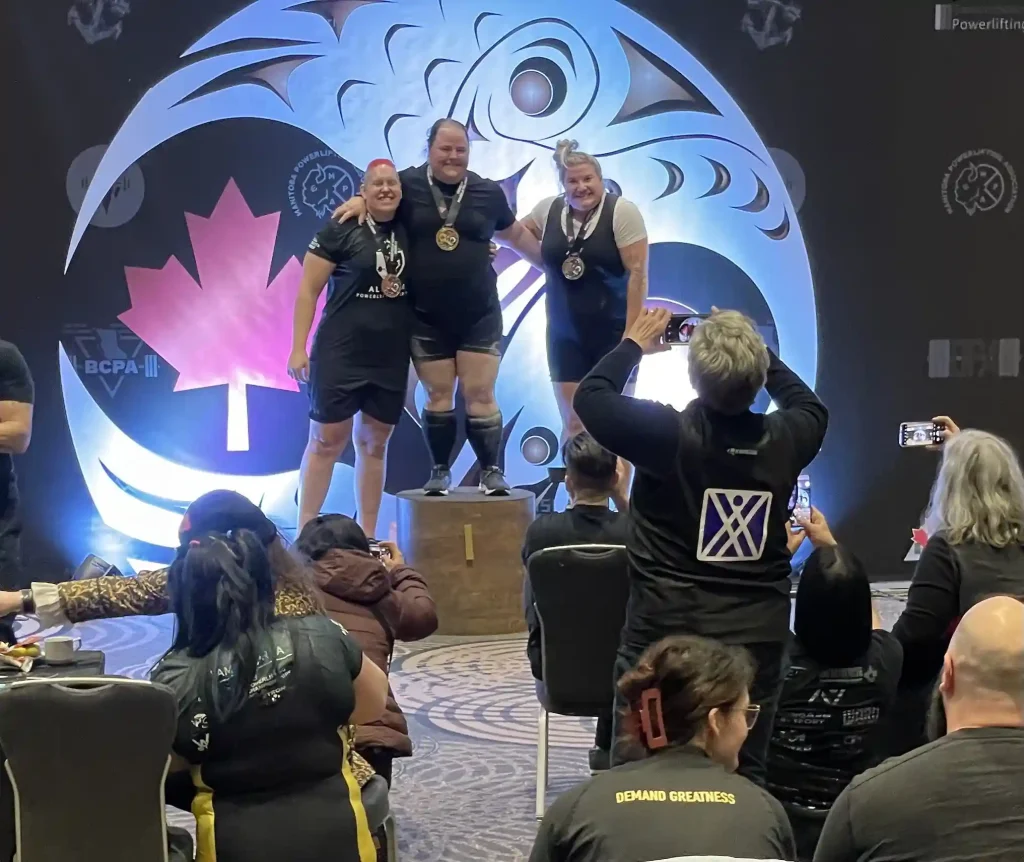 2023 CPU National Powerlifting Championships awards podium at an indoor competition venue, backed by a large circular Indigenous-style thunderbird/eagle graphic and a Canadian flag maple leaf design, with "BCPA" and other regional powerlifting association logos visible. Three medalists stand on the podium, two female, one male, wearing black competition singlets and medals around their necks. In the foreground, event attendees are seated with their backs to the camera, several wearing black jackets with the same purple "X" logo seen in earlier photos, and one shirt reads "DEMAND GREATNESS." A woman with short gray hair in the foreground holds up a phone to photograph the podium.