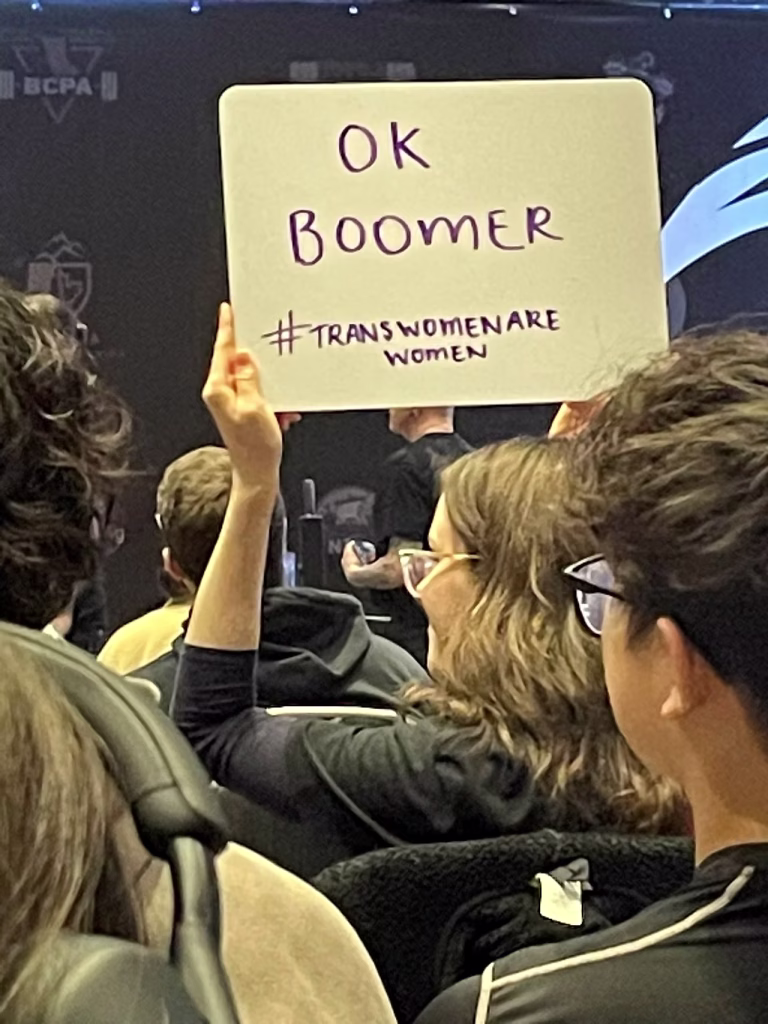  a smirking person holds up a sign that says "ok boomer #transwomenarewomen"