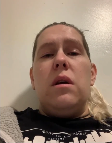 A close-up, low-angle selfie of the same man, with a somber or tearful expression, tilted head back against a plain cream-colored wall. He has shoulder-length blonde hair (visible on one side) with darker roots, and is wearing a black t-shirt reading "BAR IS LOADED" with a barbell graphic, along with a gray fuzzy garment draped over one shoulder. Small dots at the bottom of the image suggest this is a frame from a multi-slide Instagram-style post.