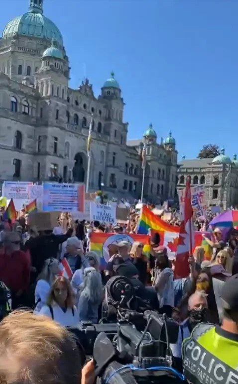 A photograph taken from within a large outdoor crowd facing toward a grand stone government building with green domed towers (the British Columbia Legislative Building in Victoria). Numerous protesters hold up rainbow Pride flags, a red Canadian flag, and various handwritten and printed signs, though the sign text is too small and distant to read clearly. A police officer wearing a high-visibility yellow vest with "POLICE" printed on the back stands in the foreground with their back to the camera, and part of a camera/media equipment setup is visible in the lower right corner.