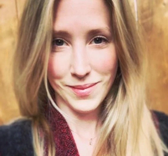 A close-up selfie photograph of a woman with long wavy blonde hair, smiling gently at the camera, wearing a dark jacket over a plaid shirt and a red patterned scarf. She stands indoors in front of a wooden slatted wall.