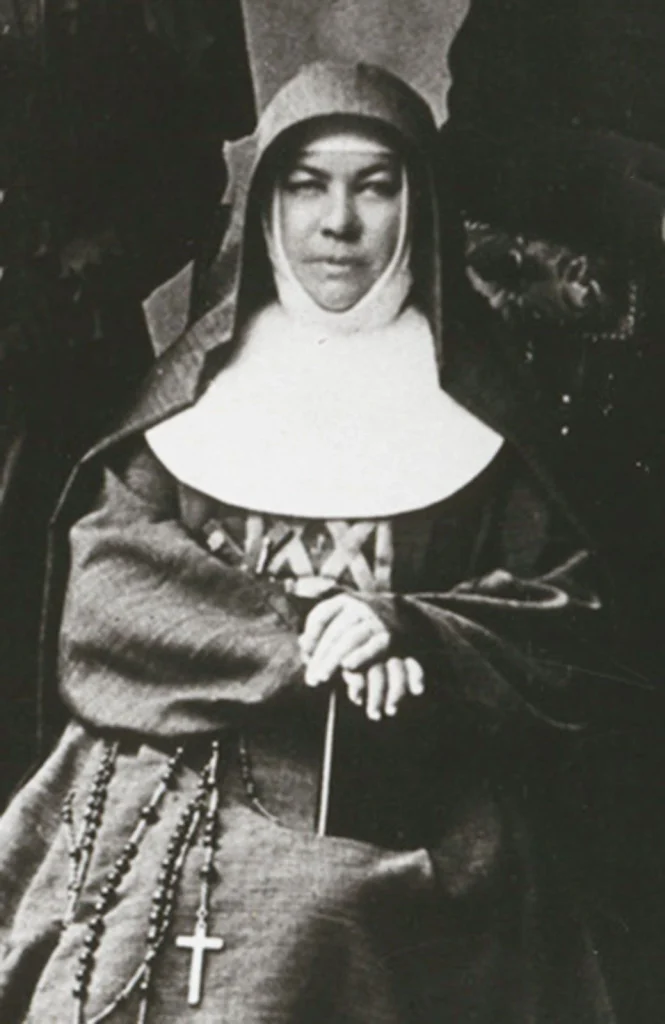 a vintage black-and-white photographic portrait of a nun in a traditional habit with a starched white wimple/collar and a tall winged coif, hands folded holding a rosary with a crucifix, several rosaries draped over her arm.