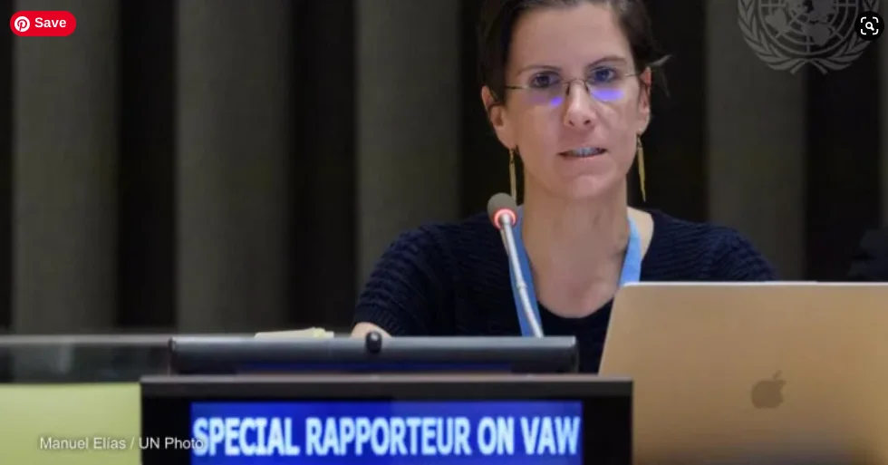 A photo of Reem Alsalem with dark hair pulled back, wearing glasses and dangling earrings, speaking into a microphone at a UN panel desk, with a laptop open in front of her. A screen or nameplate beside her reads "SPECIAL RAPPORTEUR ON VAW" in blue and white text. The UN emblem is faintly visible on the dark backdrop behind her. Photo credit in the bottom left: "Manuel Elías / UN Photo" 