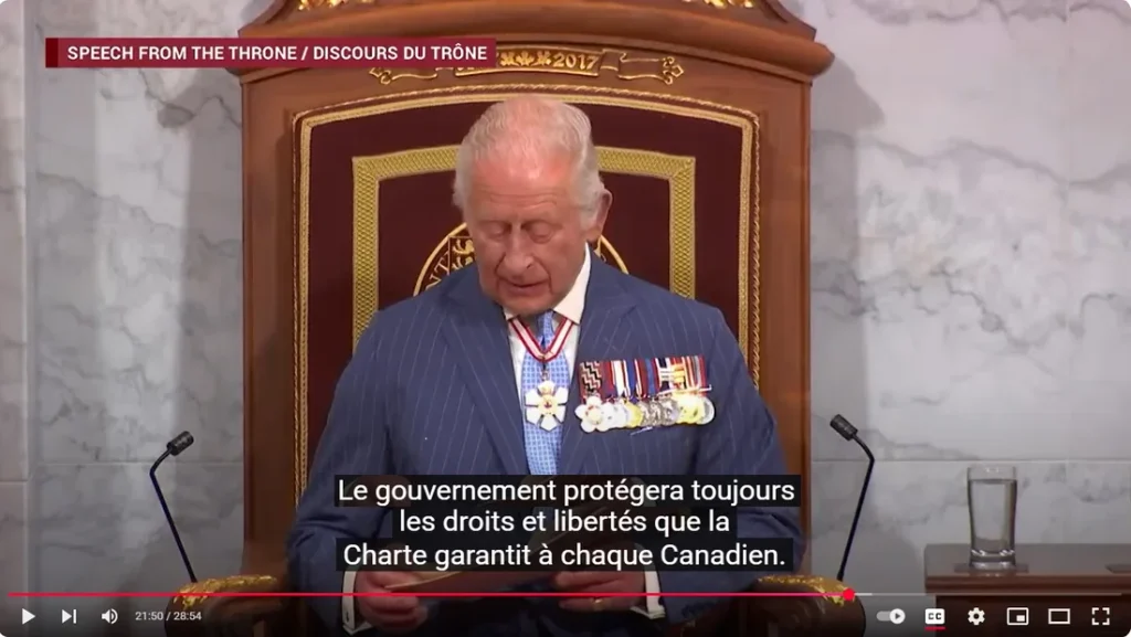 Une capture d'écran vidéo avec l'étiquette « SPEECH FROM THE THRONE / DISCOURS DU TRÔNE » et un petit filigrane « 2017 » dans la bannière supérieure. Le roi Charles III, portant un costume marine à fines rayures, une cravate bleue, une écharpe et plusieurs décorations/médailles militaires, est assis sur un trône en bois orné et lit des documents. Les sous-titres affichés en bas indiquent : « Le gouvernement protégera toujours les droits et libertés que la Charte garantit à chaque Canadien. » La barre de progression de la vidéo indique un horodatage de 21:50 sur 28:54.