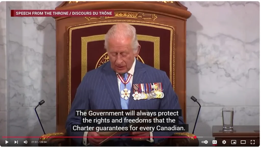 A video still labeled "SPEECH FROM THE THRONE / DISCOURS DU TRÔNE" with a small "2017" watermark graphic in the top left banner. King Charles III, wearing a pinstriped navy suit, blue tie, sash, and several military medals/honours, sits in an ornate wooden throne chair reading from papers. Subtitles overlaid at the bottom read: "The Government will always protect the rights and freedoms that the Charter guarantees for every Canadian." The video progress bar shows a timestamp of 21:51 out of 28:54.