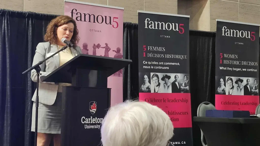 A photograph of a woman (Linda Lapointe) with curly reddish-brown hair standing at a wooden lectern reading "Carleton University," speaking into a microphone. She wears a light grey blazer over a cream-colored top and a name badge clipped to her lapel, and appears to be reading from notes on the lectern. Behind her are three vertical pink and black banners reading "famou5, OTTAWA," "5 FEMMES, 1 DÉCISION HISTORIQUE, Ce qu'elles ont commencé, nous le continuons" with black-and-white photos of women, and a partial banner with the "famou5" logo. A seated audience member with white hair is visible in the foreground, out of focus.