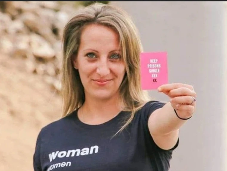 Heather Mason stands with long blonde hair smiling at the camera, wearing a black T-shirt reading "Woman" (with "women" in phonetic/IPA-style lettering beneath it), holding up a pink card that reads "KEEP PRISONS SINGLE SEX XX."