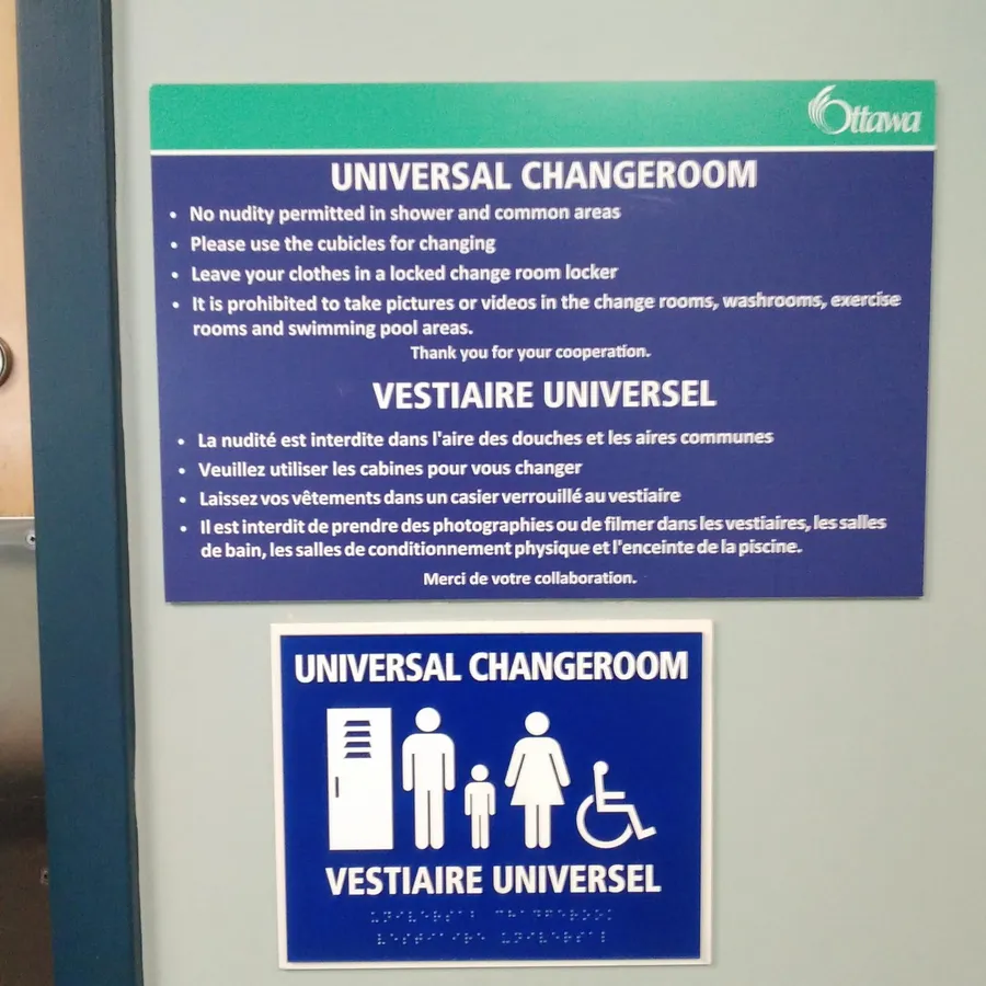 A photograph of a gray wall with two signs mounted on it, next to a partially visible door and a silver door handle/lock mechanism. The upper, larger sign has a green header bar with the City of Ottawa logo, below which bold white text on a dark blue background reads "UNIVERSAL CHANGEROOM" followed by bulleted rules: "No nudity permitted in shower and common areas," "Please use the cubicles for changing," "Leave your clothes in a locked change room locker," and "It is prohibited to take pictures or videos in the change rooms, washrooms, exercise rooms and swimming pool areas." followed by centered text "Thank you for your cooperation." Below this, the French translation appears: "VESTIAIRE UNIVERSEL" followed by "La nudité est interdite dans l'aire des douches et les aires communes," "Veuillez utiliser les cabines pour vous changer," "Laissez vos vêtements dans un casier verrouillé au vestiaire," "Il est interdit de prendre des photographies ou de filmer dans les vestiaires, les salles de bain, les salles de conditionnement physique et l'enceinte de la piscine." followed by "Merci de votre collaboration." Below this larger sign is a smaller square blue sign reading "UNIVERSAL CHANGEROOM" above a pictogram showing a locker, an adult male figure, a child figure, an adult female figure, and a wheelchair-accessibility symbol, with "VESTIAIRE UNIVERSEL" below the icons, and a line of raised Braille dots at the very bottom.