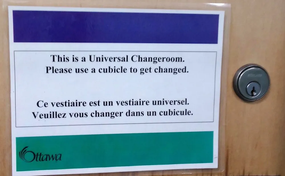 A photograph of a laminated sign affixed to a wooden door, positioned beside a metal cylindrical door lock. The sign has a dark blue header bar at the top and a teal/green footer bar at the bottom bearing the City of Ottawa logo. The white middle section contains black text reading: "This is a Universal Changeroom. / Please use a cubicle to get changed." followed by the French translation: "Ce vestiaire est un vestiaire universel. / Veuillez vous changer dans un cubicule."