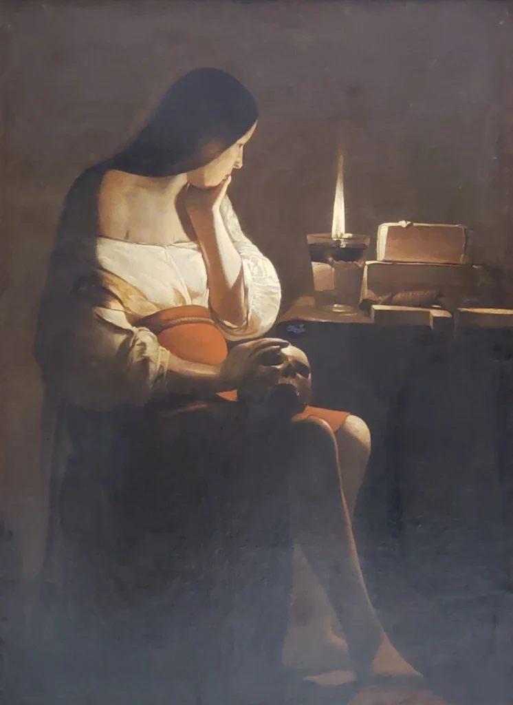 a chiaroscuro-style oil painting (Mary Magdalene by Georges de La Tour) of a woman with long dark hair, seated, resting her chin on one hand while the other rests on a skull in her lap. A single candle flame lights the scene beside a stack of books on a dark table.