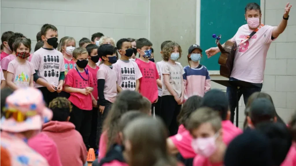 A photo inside a school gymnasium showing a large group of children, most wearing pink shirts and protective face masks, standing in rows facing a man on the right who is playing an acoustic guitar and raising one hand while singing; he wears a pink polo shirt and has a name badge/lanyard. More children sit facing him in the blurred foreground. Visible shirt text includes: "I Celebrate Diversity DAY OF PINK" (with five stick-figure icons), "A PINK SHIRT CAN STOP HATE," and "KIND HUMAN BE[ING]."