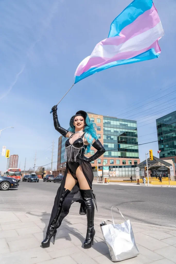 A photo of a drag queen performer with long blue hair, standing on a city sidewalk holding up a trans pride flag (light blue, pink, white, pink, light blue stripes) attached to a pole. He's wearing a black outfit with a fringed metal-chain bodice, a small black cap, thigh-high black boots, and a sheer skirt panel with a high slit, one hand on his hip. A silver tote bag sits on the ground beside him; office buildings and street traffic are visible behind him.