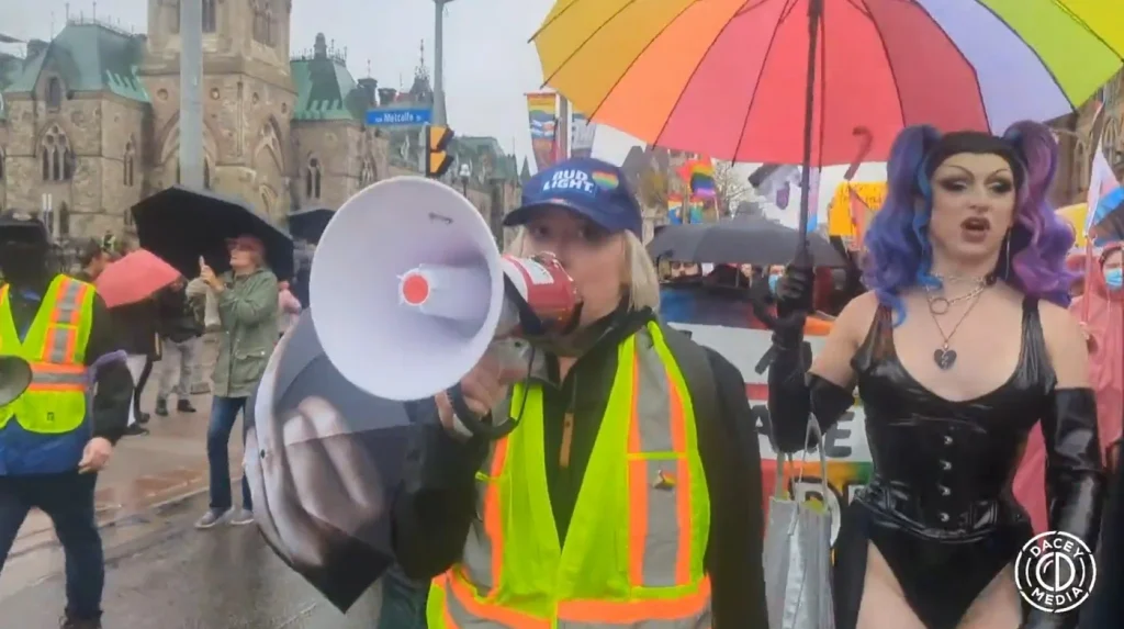A video still from a pride parade on a rainy day. In the foreground, a woman in a blue "BUD LIGHT" cap and a yellow safety vest speaks into a red-and-white megaphone. To her right, a male drag performer with purple-and-blue pigtails, heavy makeup, a black vinyl corset top and jewelry holds a large rainbow-and-trans-flag-colored umbrella. Other people with umbrellas and rainbow flags are visible in the background, along with a stone government building. A "DACEY MEDIA" logo watermark appears in the bottom right corner.